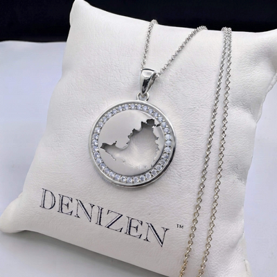A silver-tone oval pendant necklace with a cutout of St. Maarten map and a halo of clear stones, displayed on a white cushion printed with the brand name DENIZEN.