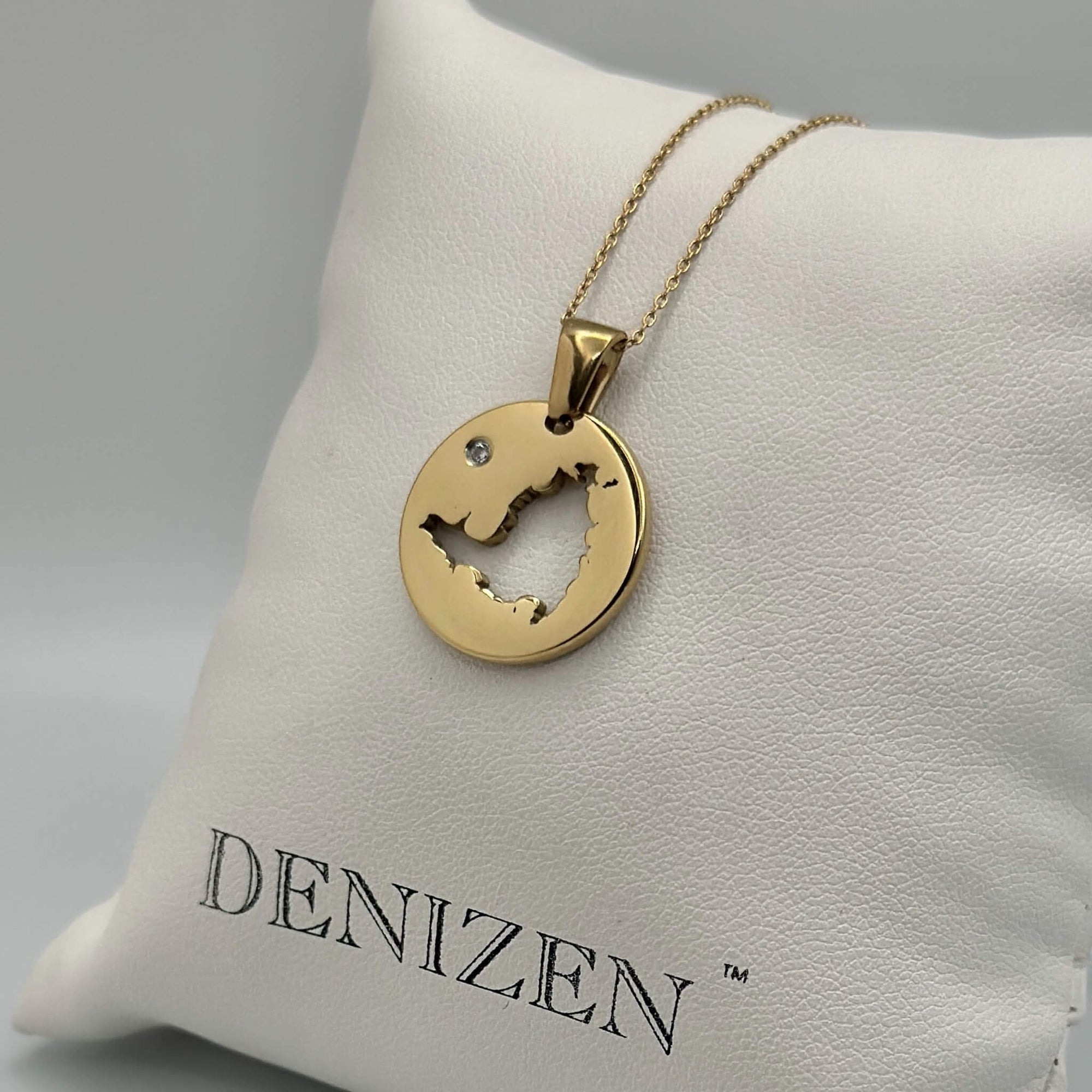 A round gold pendant necklace with a cutout of St. Maarten map an a small clear stone resting on a white textured cushion printed with the brand name 'DENIZEN'.