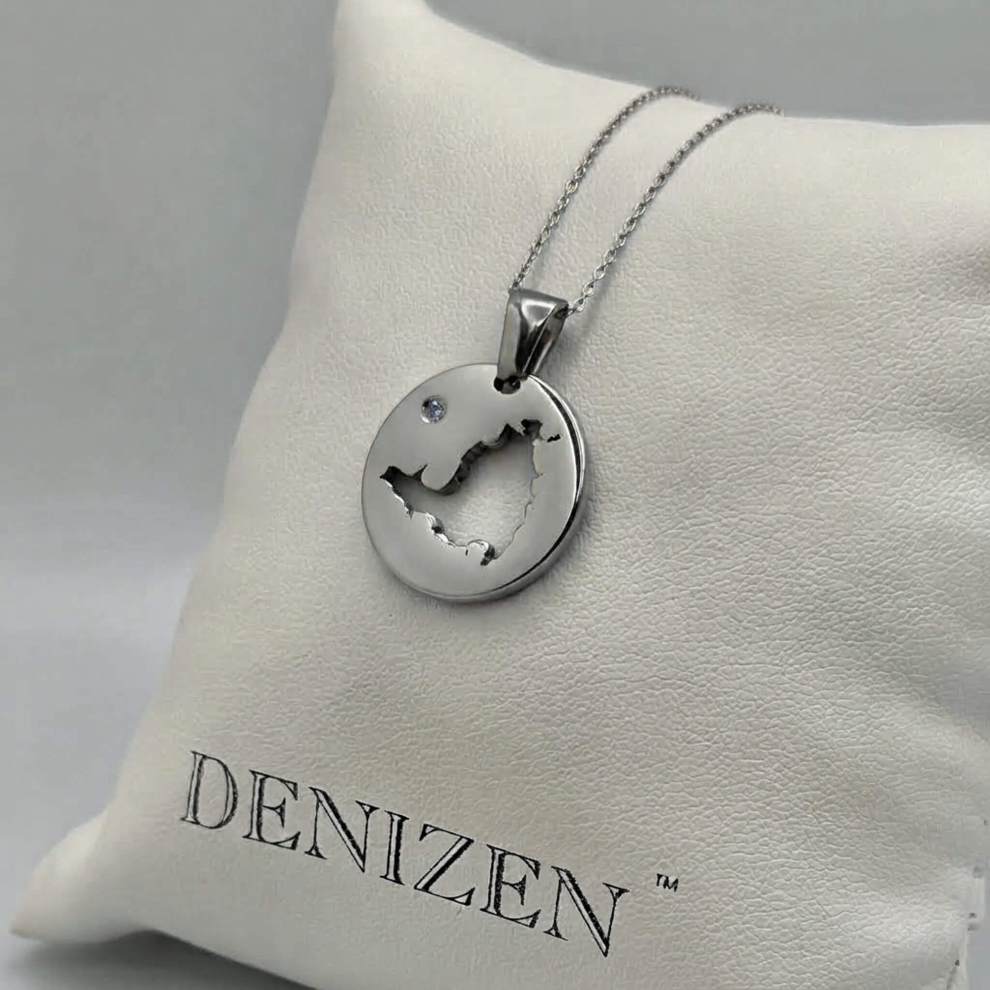 A silver-toned round pendant necklace with a cutout of the map of St. Maarten and a small clear stone, displayed on a white cushion branded DENIZEN.