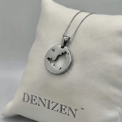 A silver-toned round pendant necklace with a cutout of the map of St. Maarten and a small clear stone, displayed on a white cushion branded DENIZEN.