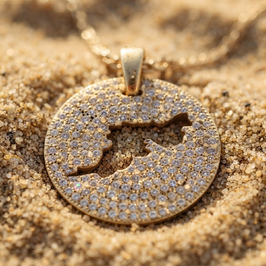 A gold round pendant necklace covered in diamond pavé, featuring a cut-out map of Tortola in the center, resting on sand.