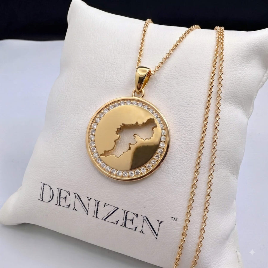 A gold round pendant necklace covered in diamond pavé, featuring a cut-out map of Tortola in the center, resting on sand.
