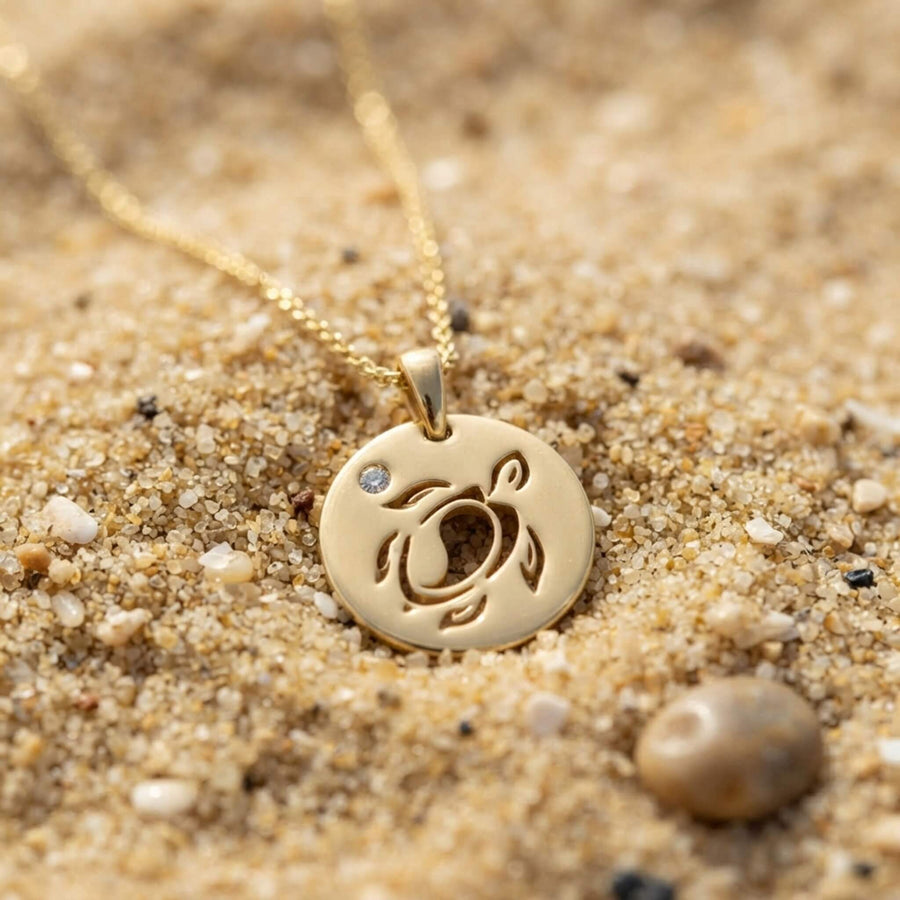 A gold-toned round pendant necklace with a cutout sea turtle design and a small clear stone, resting on sandy beach ground.