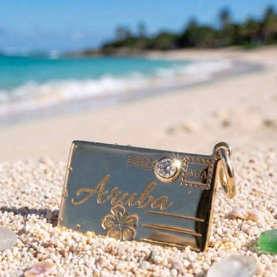 A gold postcard charm engraved with 'Aruba,' an hibiscus motif, and a small crystal, resting on white sand with a blurred beach and turquoise water in the background.