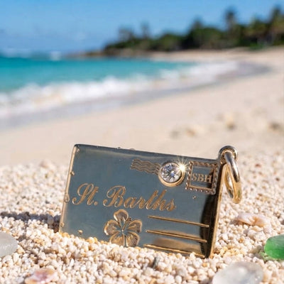 A gold postcard charm engraved with 'St Barths,' an hibiscus motif, and a small crystal resting on white sand with a blurred beach and turquoise water in the background.