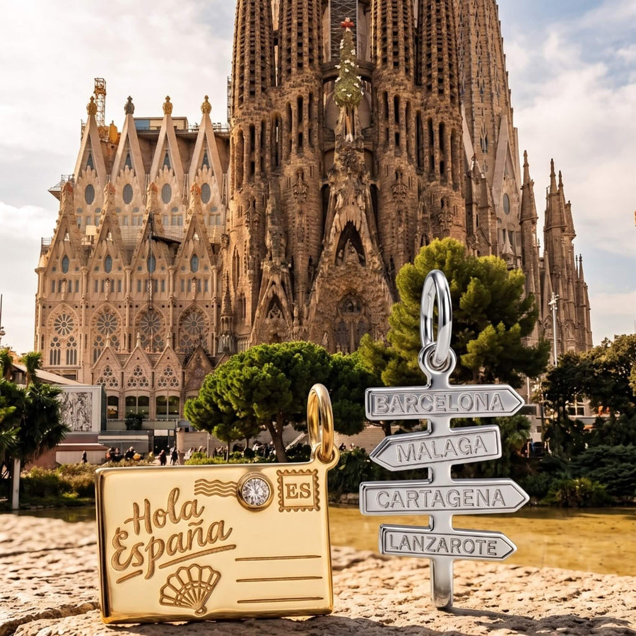 A silver signpost charm and a gold postcard charm displayed outdoors in front of Sagrada Familia in Barcelona. 