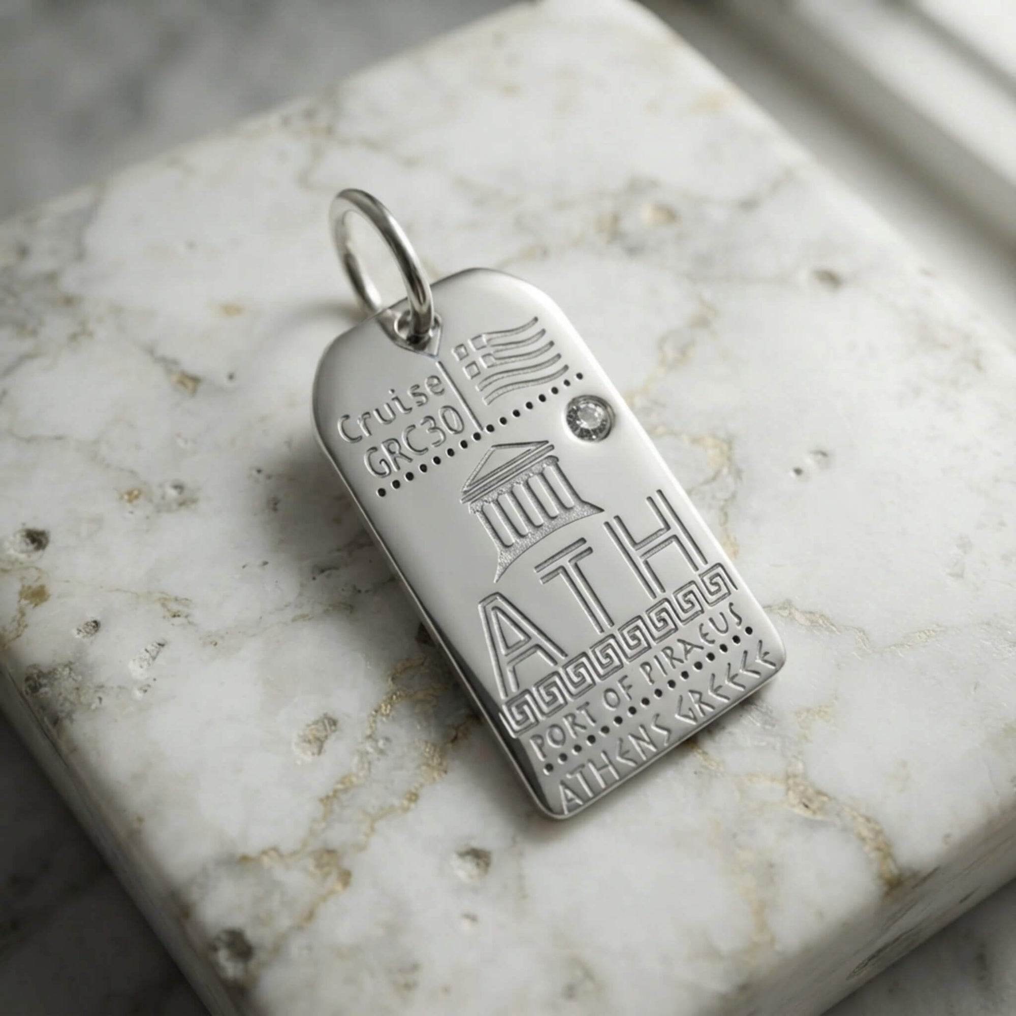 A silver tag charm engraved with Athens airport code and details such as the Acropolis and Greek flag, resting on a white marble slab.