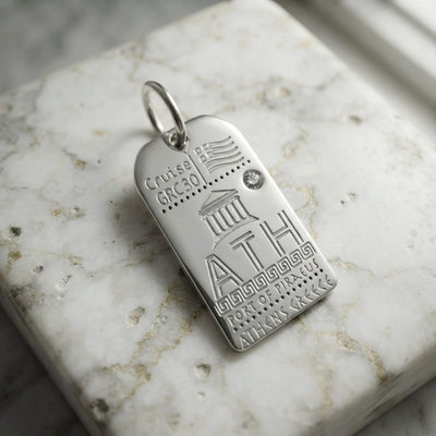 A silver tag charm engraved with Athens airport code and details such as the Acropolis and Greek flag, resting on a white marble slab.