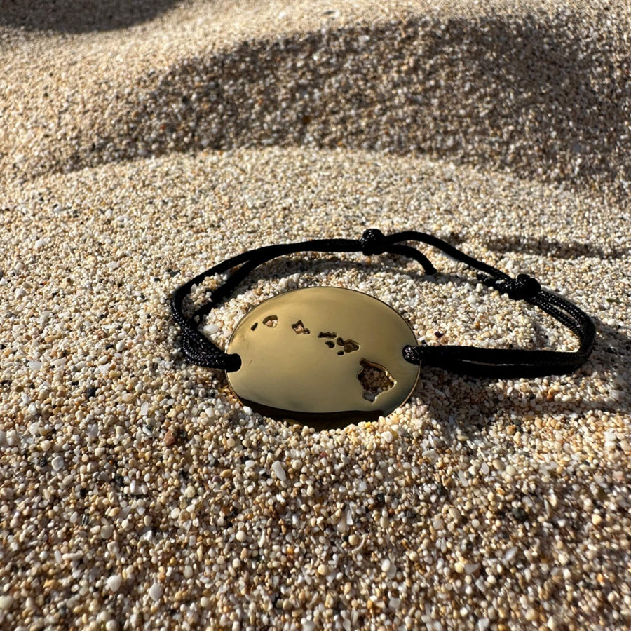 A rose gold pendant necklace with a cutout map of Hawaii and a small clear gemstone, displayed on a sandy beach with a pink hibiscus flower, green leaves, and lava rocks.